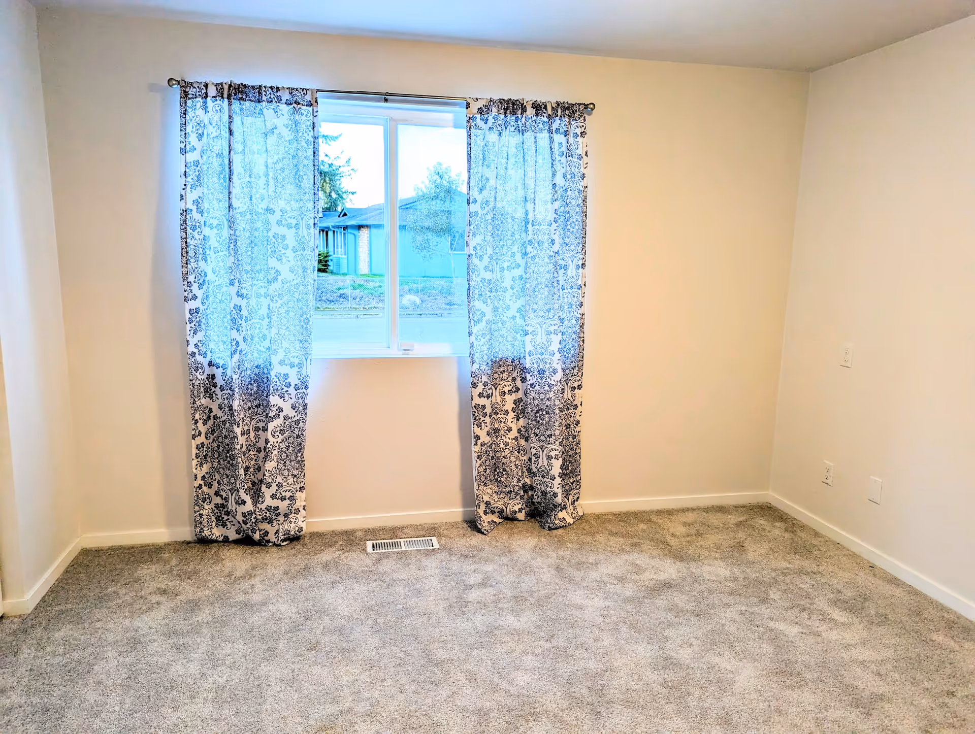 Empty room with light gray carpet, beige walls, and a window with patterned blue and white curtains. Outside the window, a street and neighboring houses are visible.