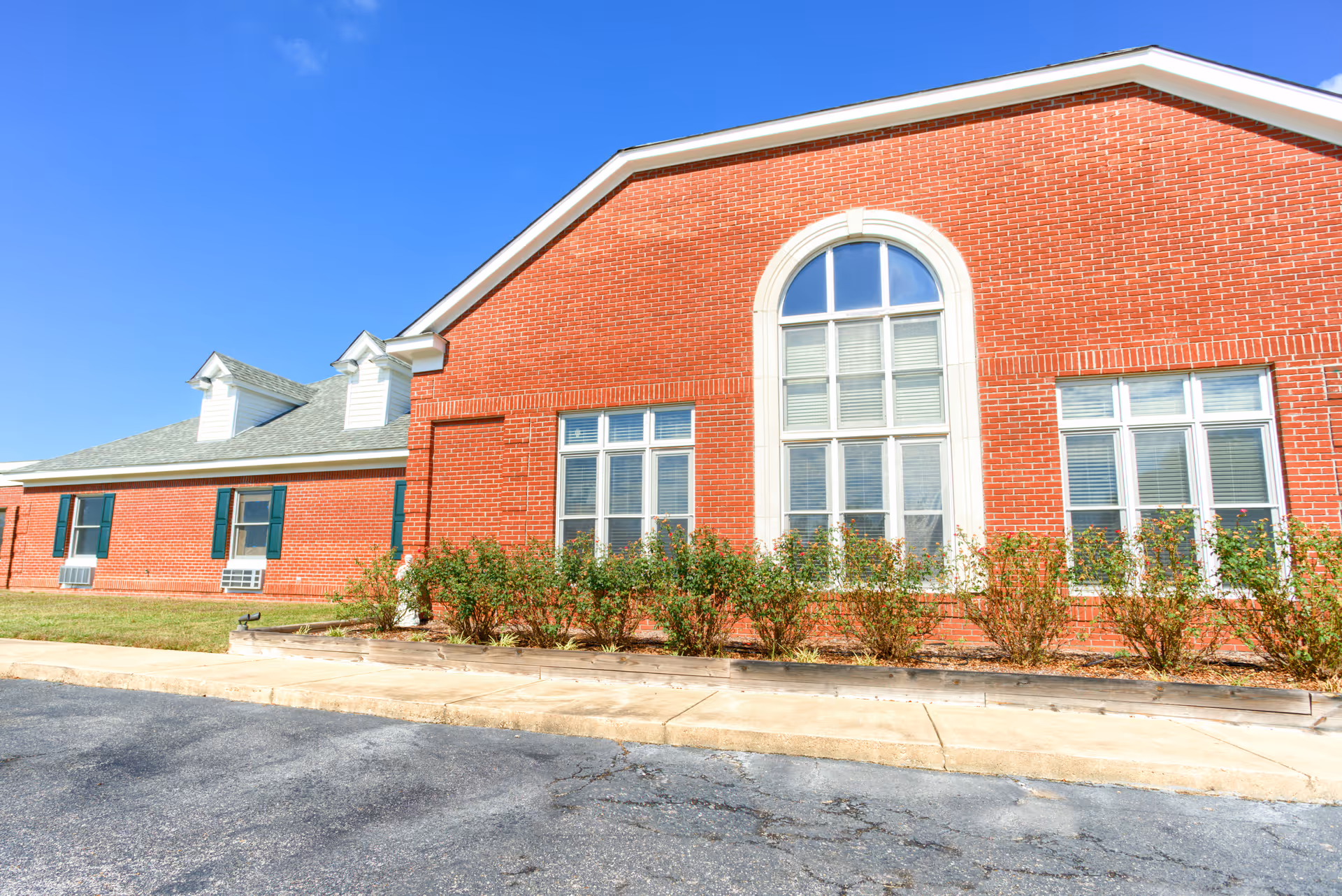 Exterior view of a red brick building with multiple windows, green shutters, and a gray shingled roof under a clear blue sky. There are small bushes planted along the building and a paved driveway in front.