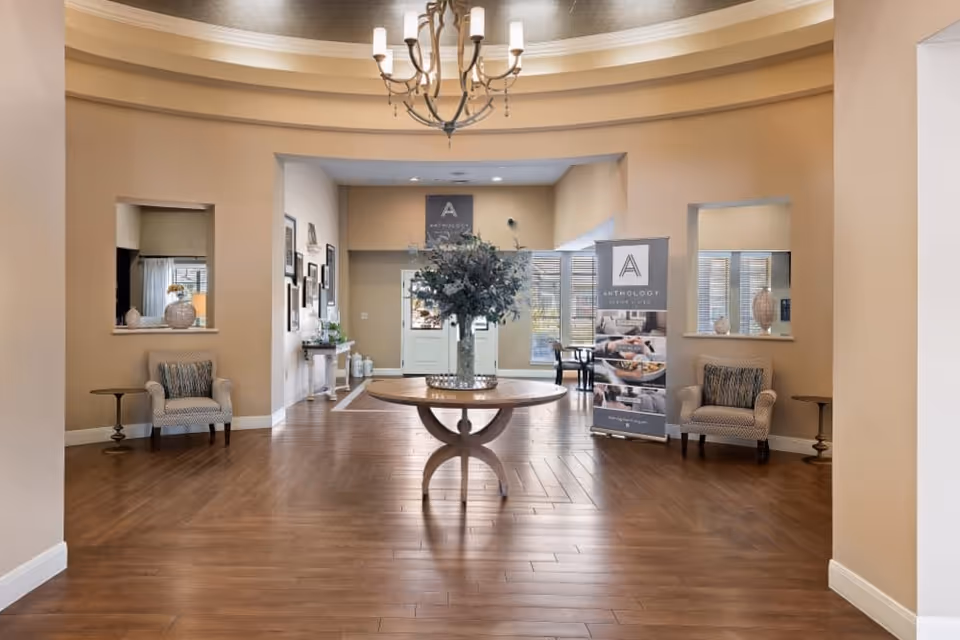Spacious and well-lit interior lobby area with wooden flooring, a round wooden table with a large floral arrangement in the center, two armchairs with patterned cushions on either side, small side tables, and a chandelier hanging from the ceiling. There are promotional banners for Anthology Senior Living near the back entrance with large windows allowing natural light to enter.