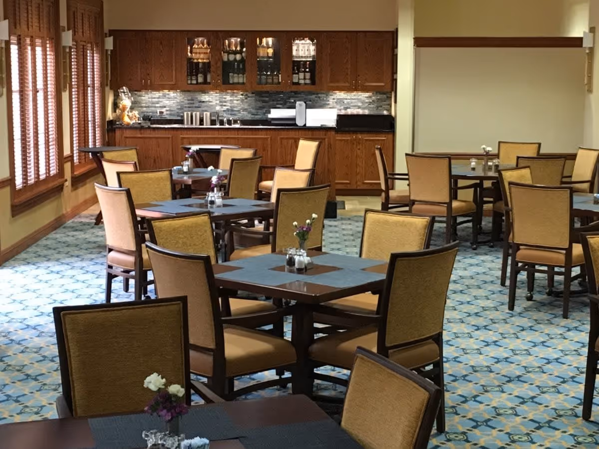A dining room with multiple square tables and cushioned chairs arranged neatly. Each table has small flower vases and salt and pepper shakers. The room features large windows with wooden blinds on the left side and a wooden cabinet with glass doors and a countertop along the back wall.
