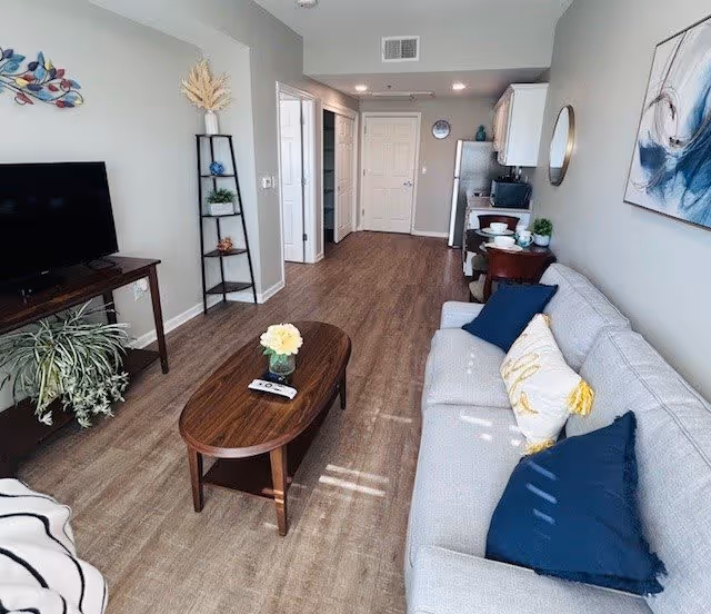 A modern living room with a light gray sofa adorned with blue and white pillows, a wooden coffee table with a flower vase and remote controls, a TV on a wooden stand, a black metal shelf with decorative items, and a kitchen area in the background with white cabinets and a refrigerator.
