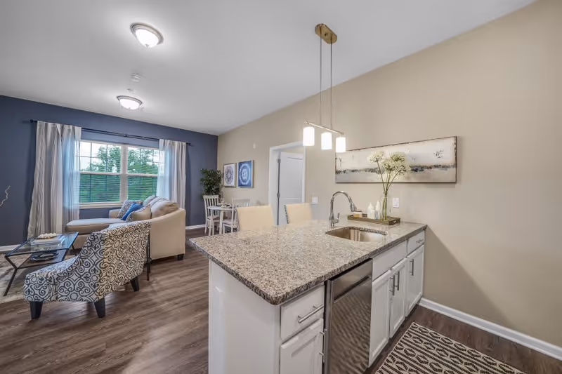 A modern open-concept living space featuring a kitchen island with granite countertop and sink, two beige chairs, and pendant lighting. In the background, there is a living room area with a patterned armchair, beige sofa, glass coffee table, and a large window with curtains. A small dining table with chairs and wall art is also visible.