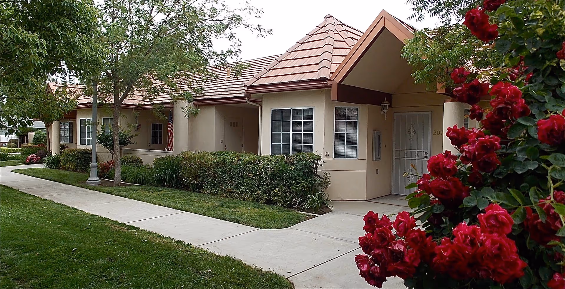 Exterior view of a single-story building with beige walls and a tiled roof, surrounded by green shrubs, trees, and vibrant red flowers in the foreground. A concrete sidewalk runs alongside the building, and an American flag is visible near one of the entrances.