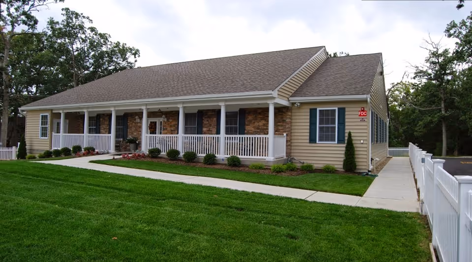 Single-story beige building with a covered front porch, white railing, walkway, and manicured lawn.