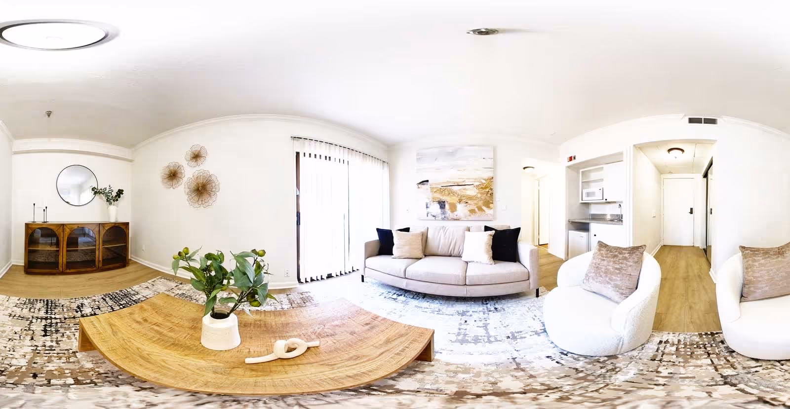 Bright and modern living room with a beige sofa adorned with black and beige pillows, two white cushioned armchairs with beige pillows, a wooden coffee table with a small plant and decorative item, a patterned area rug, a wooden cabinet with a round mirror and vase with greenery, wall art, and vertical blinds covering a large window or sliding door.