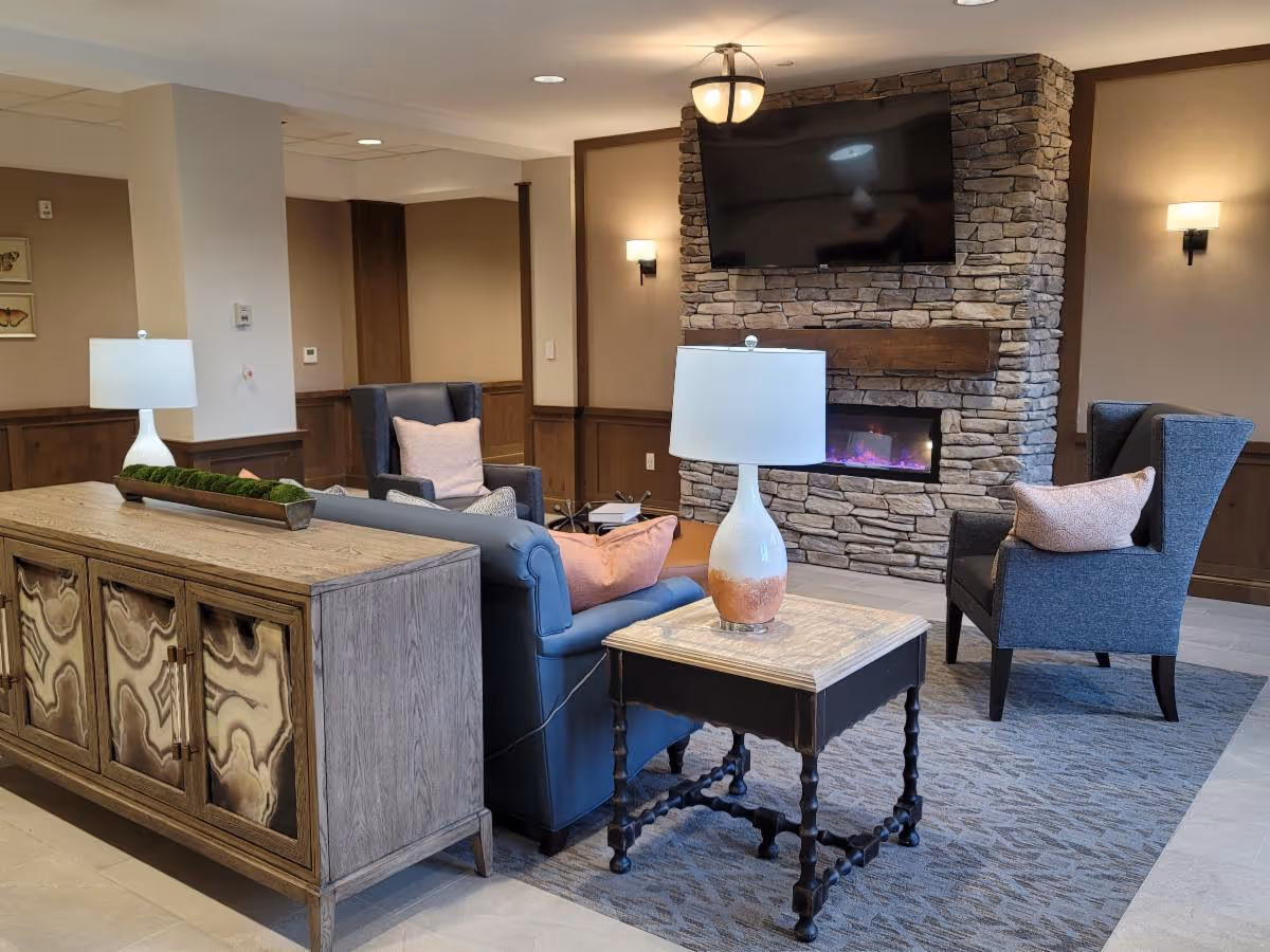A cozy living room area featuring a stone fireplace with a mounted flat-screen TV above it. The room includes a blue sofa with pink pillows, two gray armchairs with pink cushions, a wooden side table with a white and orange lamp, and a wooden cabinet with decorative panels. The walls have wood paneling and are painted in neutral tones, with wall sconces providing additional lighting.