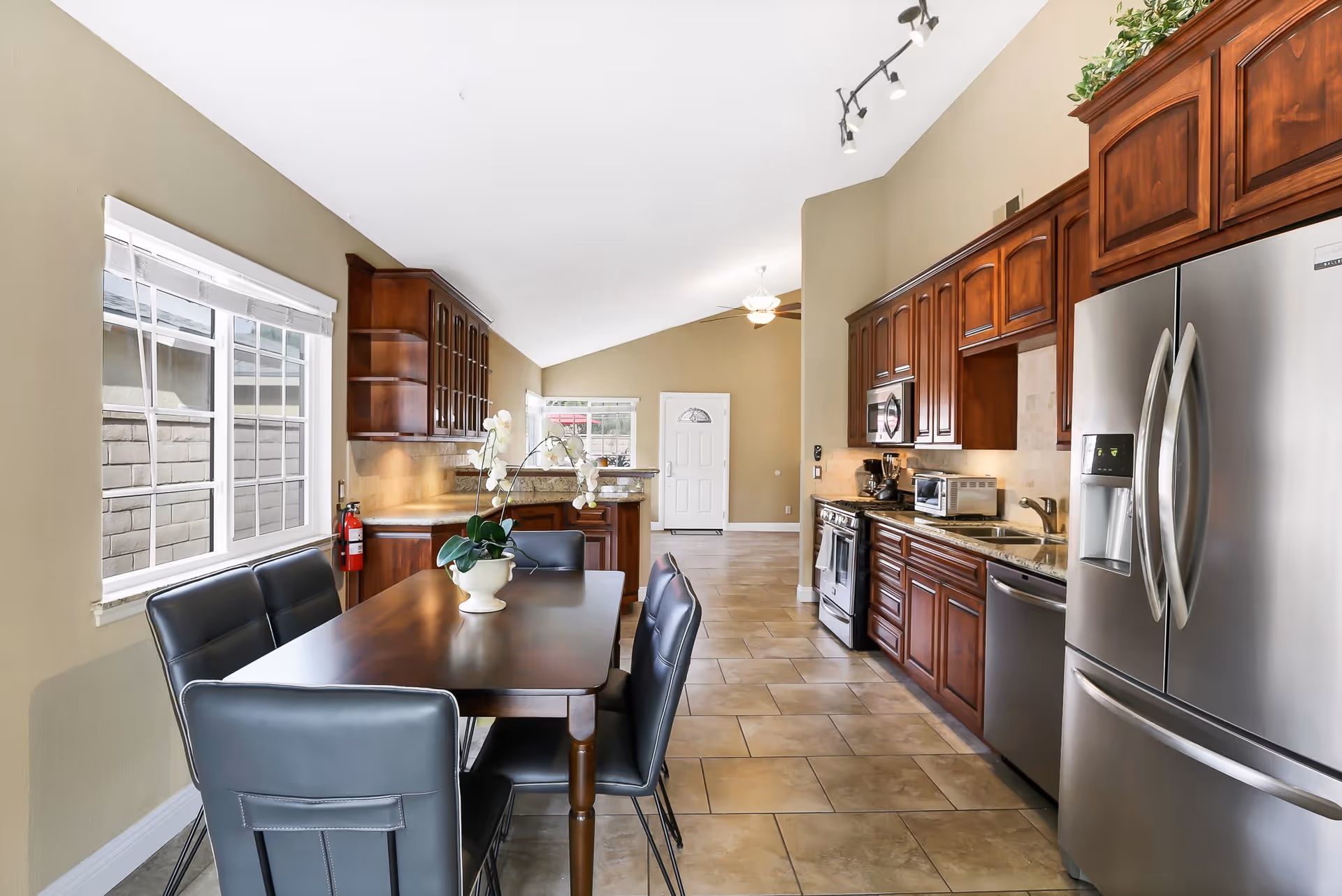 A bright kitchen and dining area with beige walls and tiled floor. The kitchen features dark wood cabinets, stainless steel refrigerator, dishwasher, stove, microwave, and a toaster oven. A dark wood dining table with six black leather chairs is positioned near a window with white blinds. A white orchid plant is placed on the dining table. The ceiling is vaulted with track lighting and a ceiling fan.