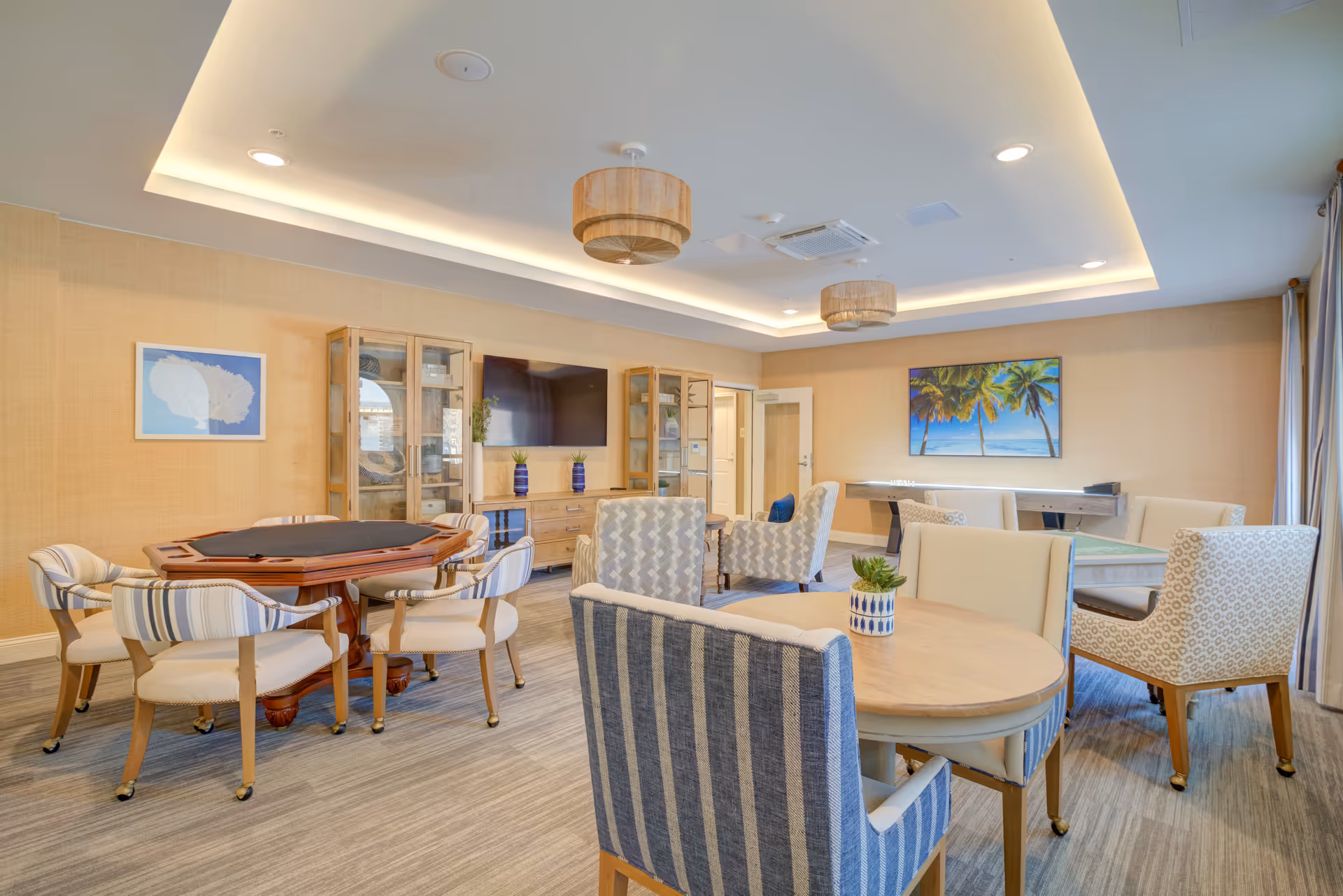 Bright, well-lit senior living common room with card tables, upholstered chairs, a TV, and coastal artwork on the walls.