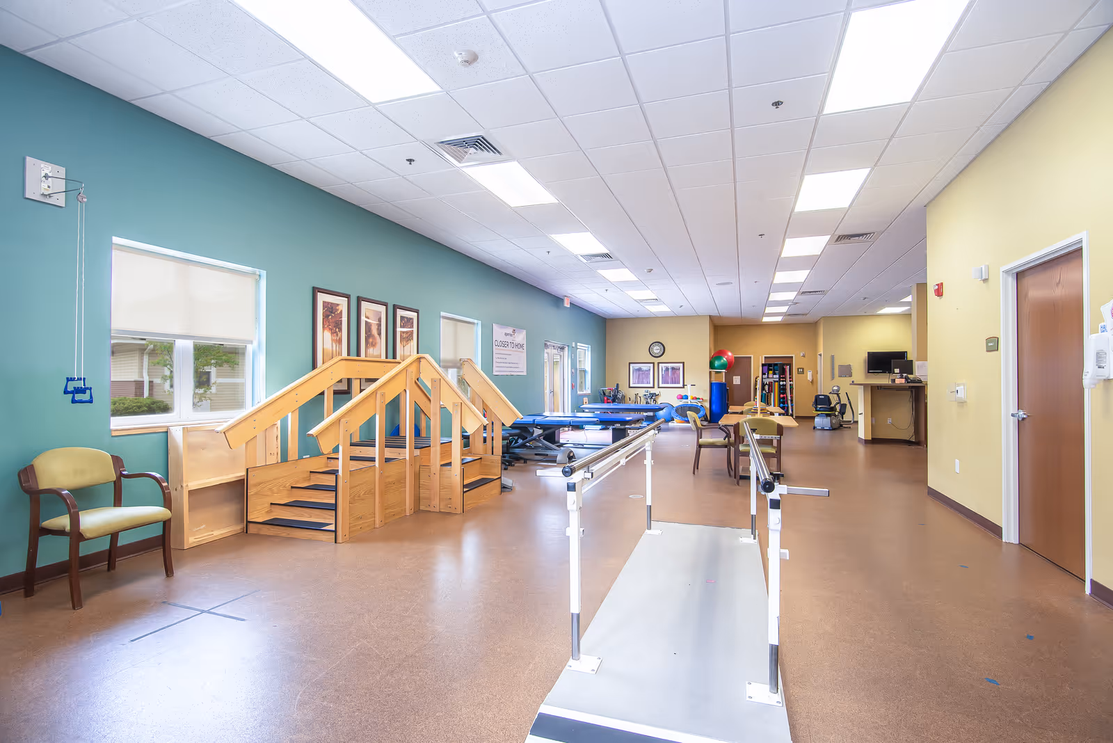 A spacious rehabilitation or physical therapy room with wooden stairs for practice, parallel bars for walking exercises, chairs, exercise balls, and therapy tables. The walls are painted green and yellow, and the room is well-lit with ceiling lights and windows.