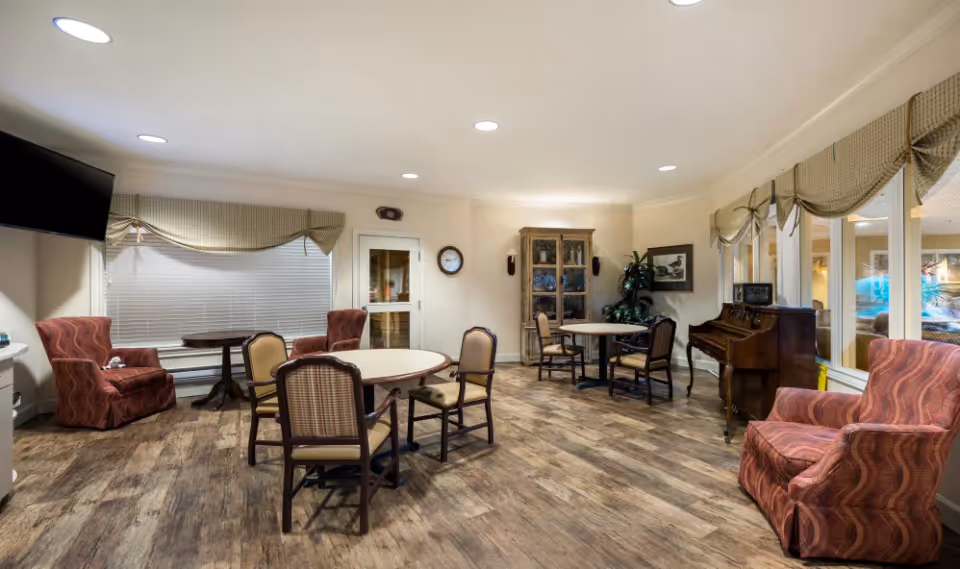 A cozy common area in a senior living facility featuring round tables with chairs, several armchairs with patterned upholstery, a wooden piano, a cabinet with glass doors, and large windows with valance curtains. The room has wood-style flooring and recessed ceiling lights.