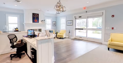 Bright and spacious assisted living facility lobby with a reception desk on the left, office chair, computer monitors, and paperwork. The room features light-colored walls, large windows with white blinds, a fireplace with artwork above it, two yellow armchairs, and a glass door leading outside.