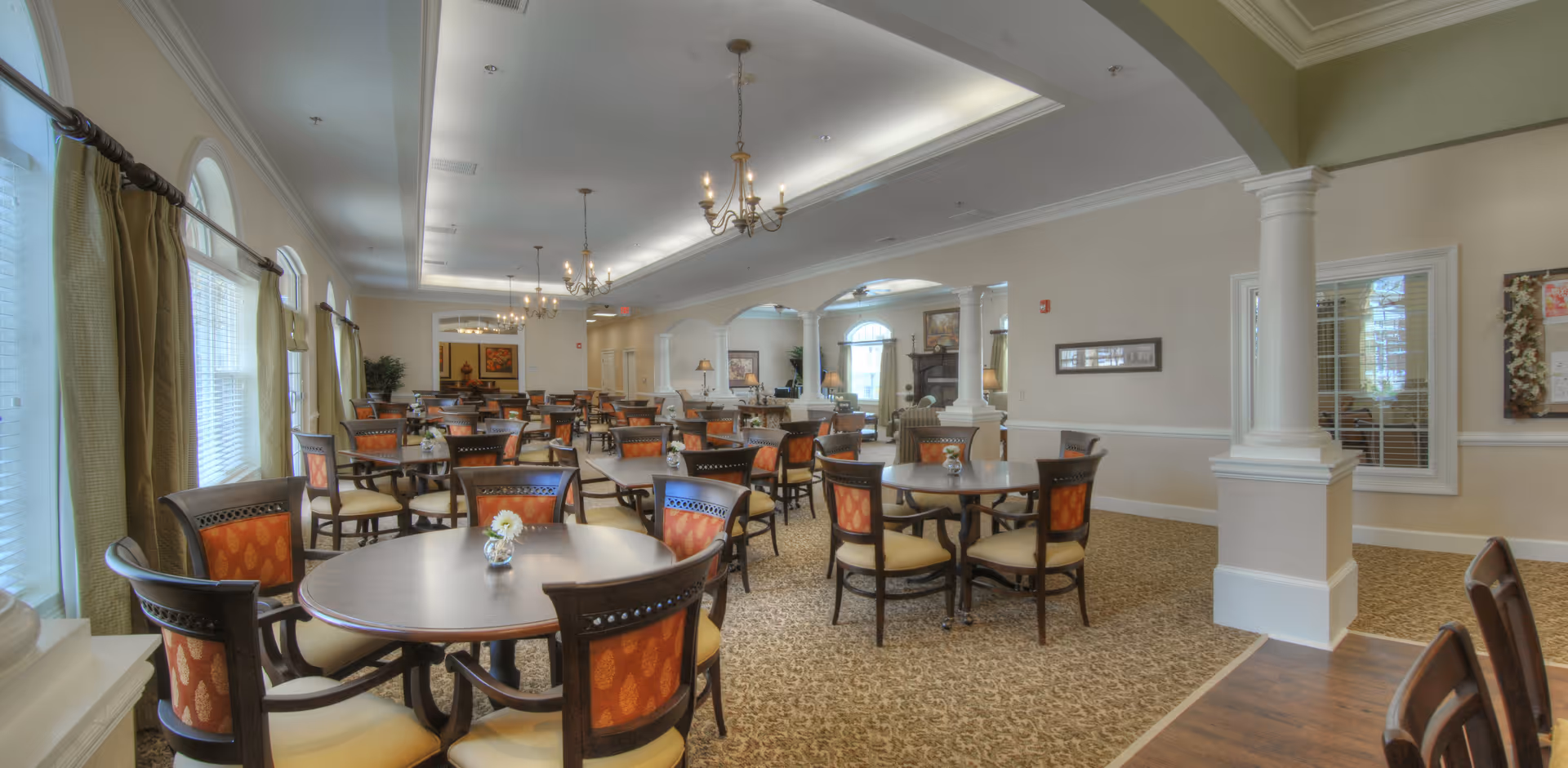 Spacious dining room with multiple round tables and chairs, chandeliers, and large arched windows.