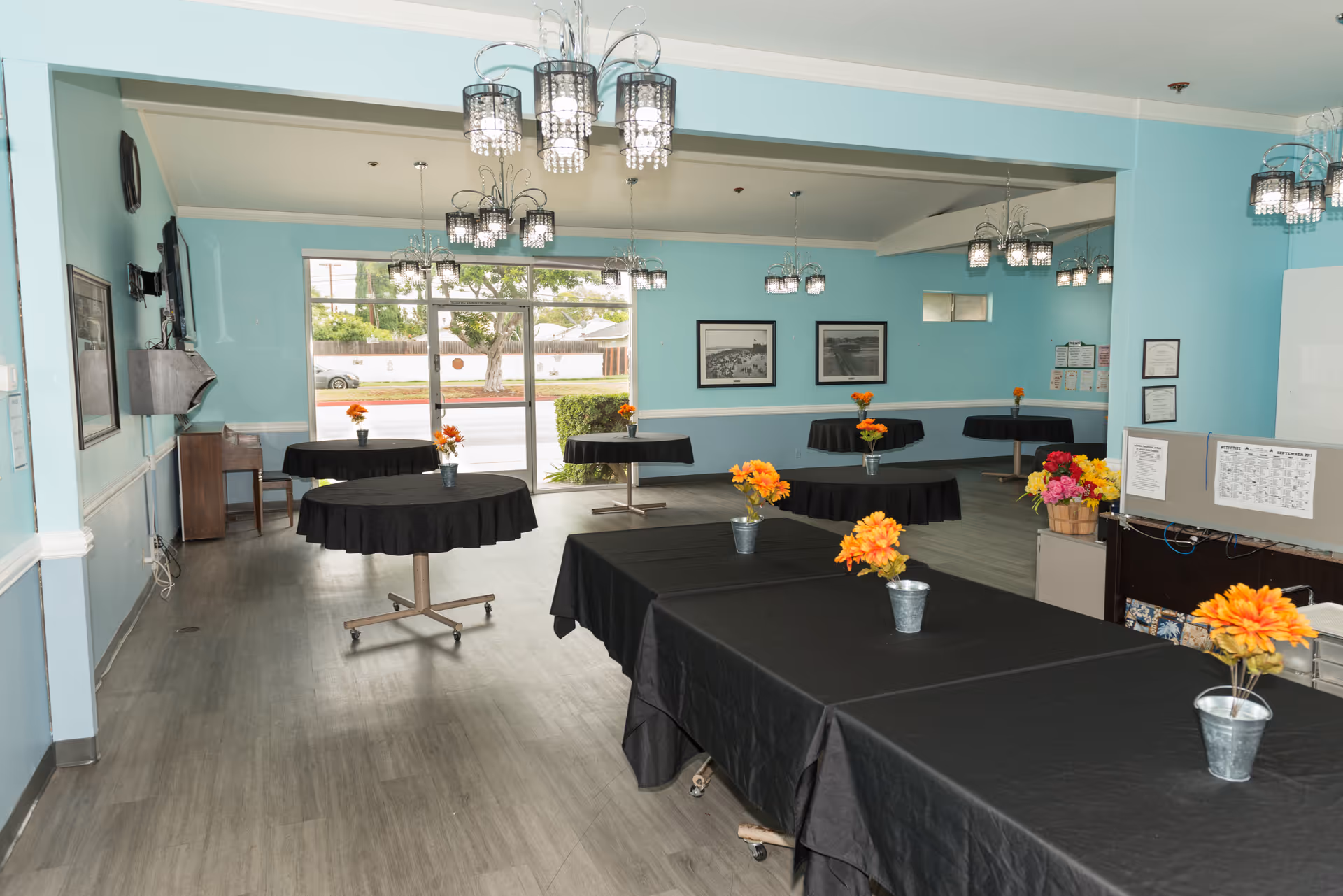 Interior view of a dining room with light blue walls and multiple round and rectangular tables covered with black tablecloths. Each table has a small metal bucket with orange flowers. The room has several modern chandeliers hanging from the ceiling and large windows letting in natural light. There are framed pictures on the walls and a small desk area with flowers and papers on the right side.