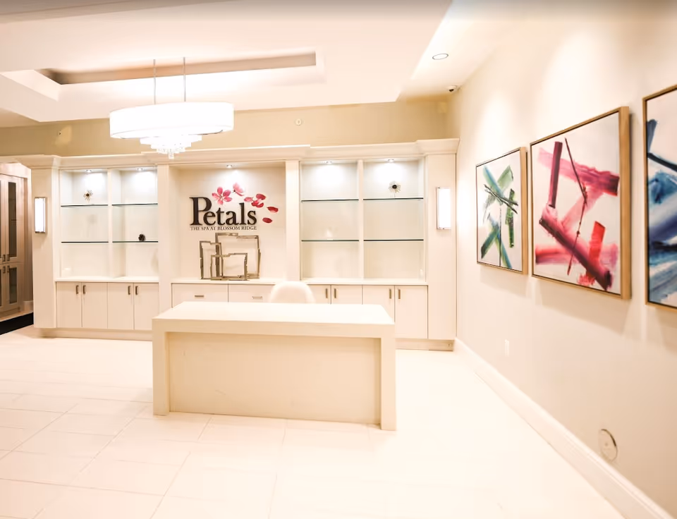 A modern spa reception area with a white desk in the center, built-in shelves with decorative items, and a sign that reads 'Petals The Spa at Blossom Ridge'. The walls are light-colored and adorned with three abstract paintings. A large, round ceiling light fixture hangs above the desk.