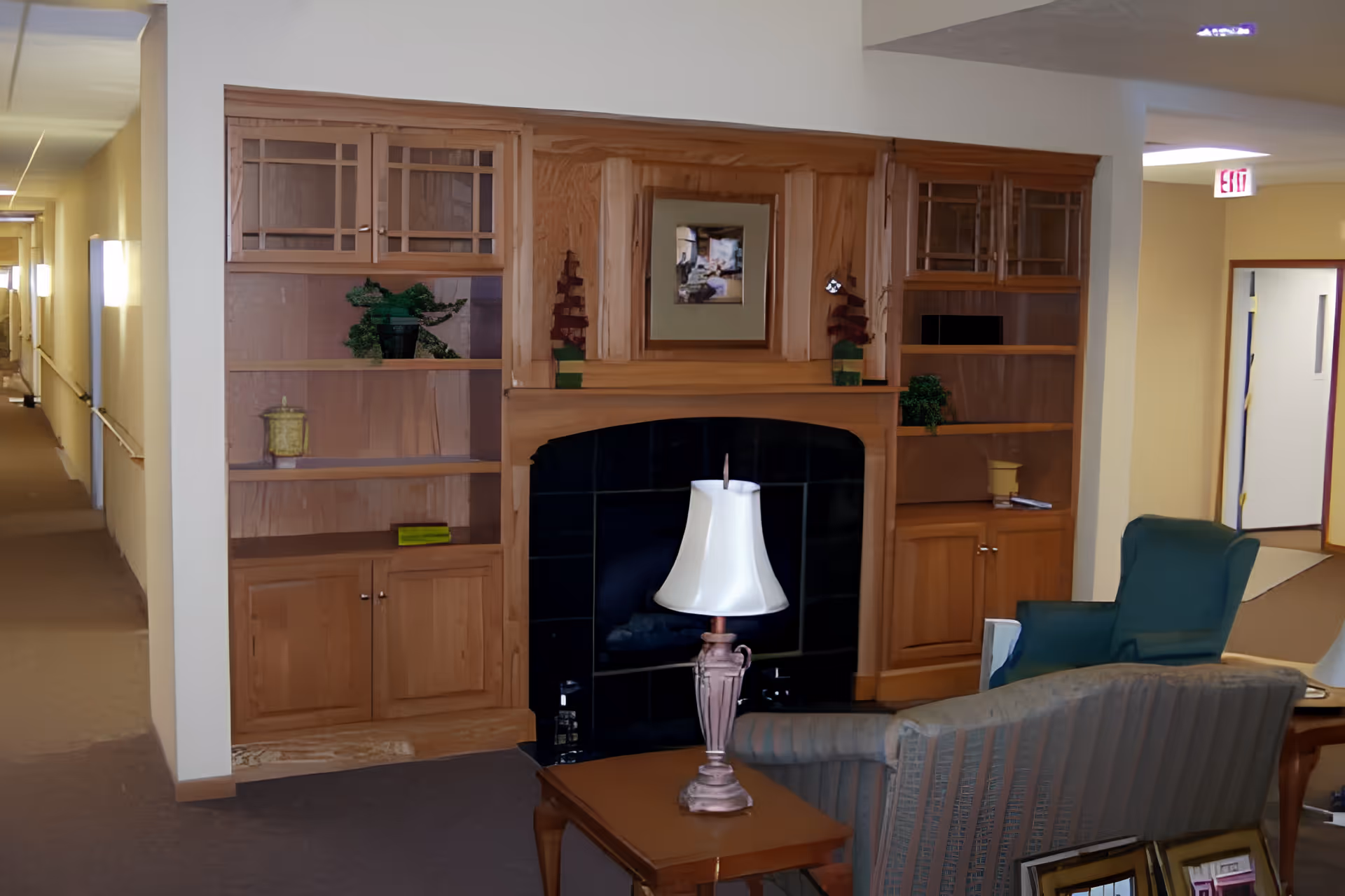 A cozy interior common area with a wooden built-in shelving unit and fireplace. The shelves hold decorative items including small plants and a framed picture above the fireplace. In front of the fireplace is a wooden table with a lamp, and there are upholstered chairs nearby. A hallway extends to the left with wall-mounted lights and handrails.