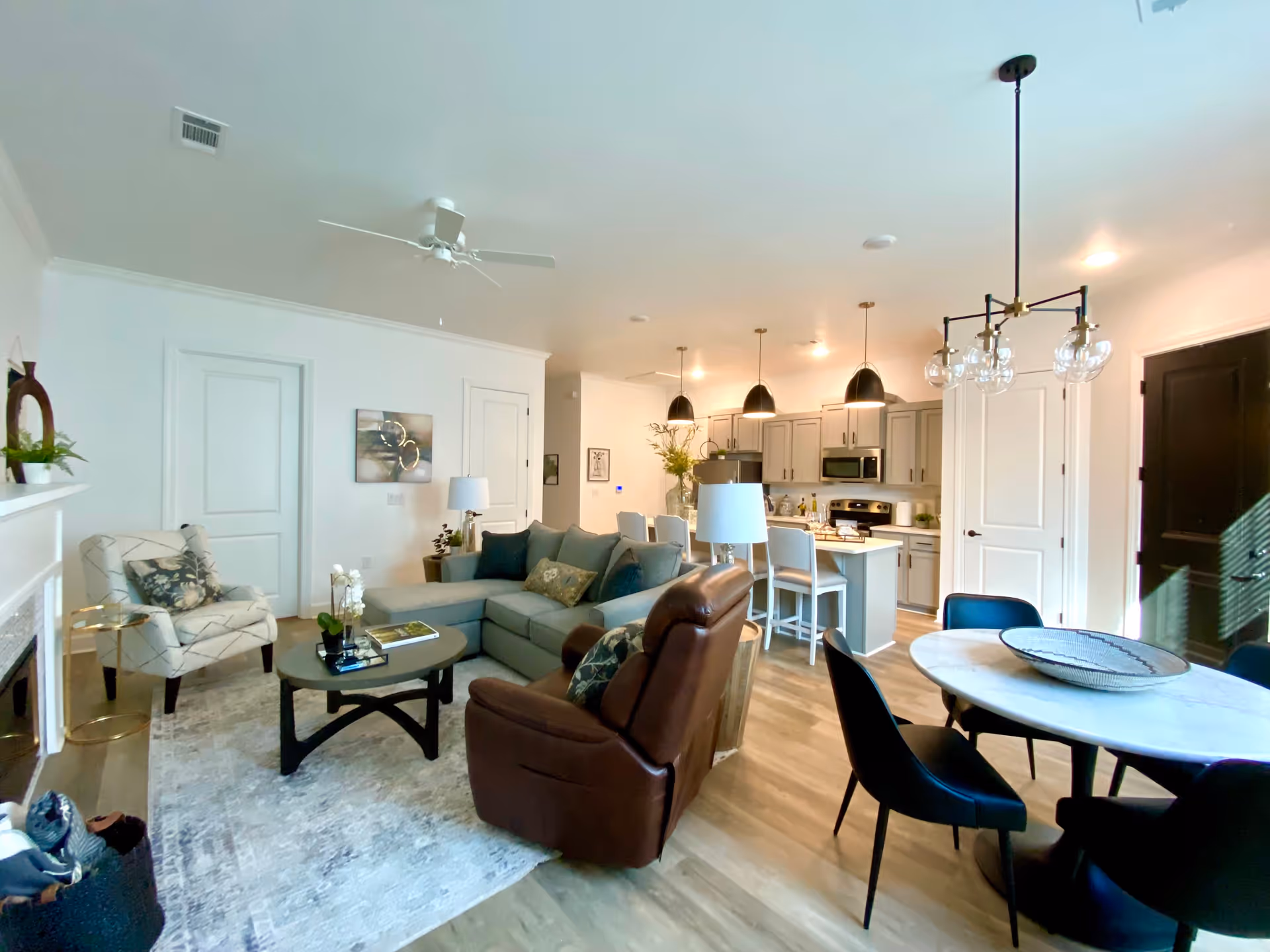 A bright and modern open-concept living area featuring a gray sectional sofa, a brown recliner, and a patterned armchair arranged around a coffee table on a light-colored rug. The space includes a kitchen with light gray cabinets, stainless steel appliances, and a kitchen island with bar stools. A round dining table with black chairs is also visible, along with contemporary light fixtures and wood flooring.