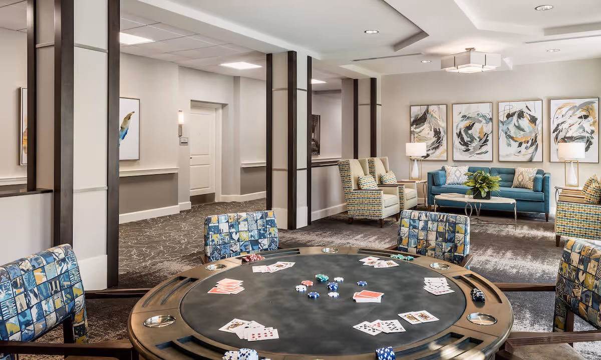 A spacious and well-lit common area in a senior living facility featuring a round poker table with playing cards and poker chips, surrounded by patterned chairs. In the background, there are comfortable armchairs and a teal sofa with decorative pillows, abstract artwork on the wall, and two table lamps providing warm lighting.