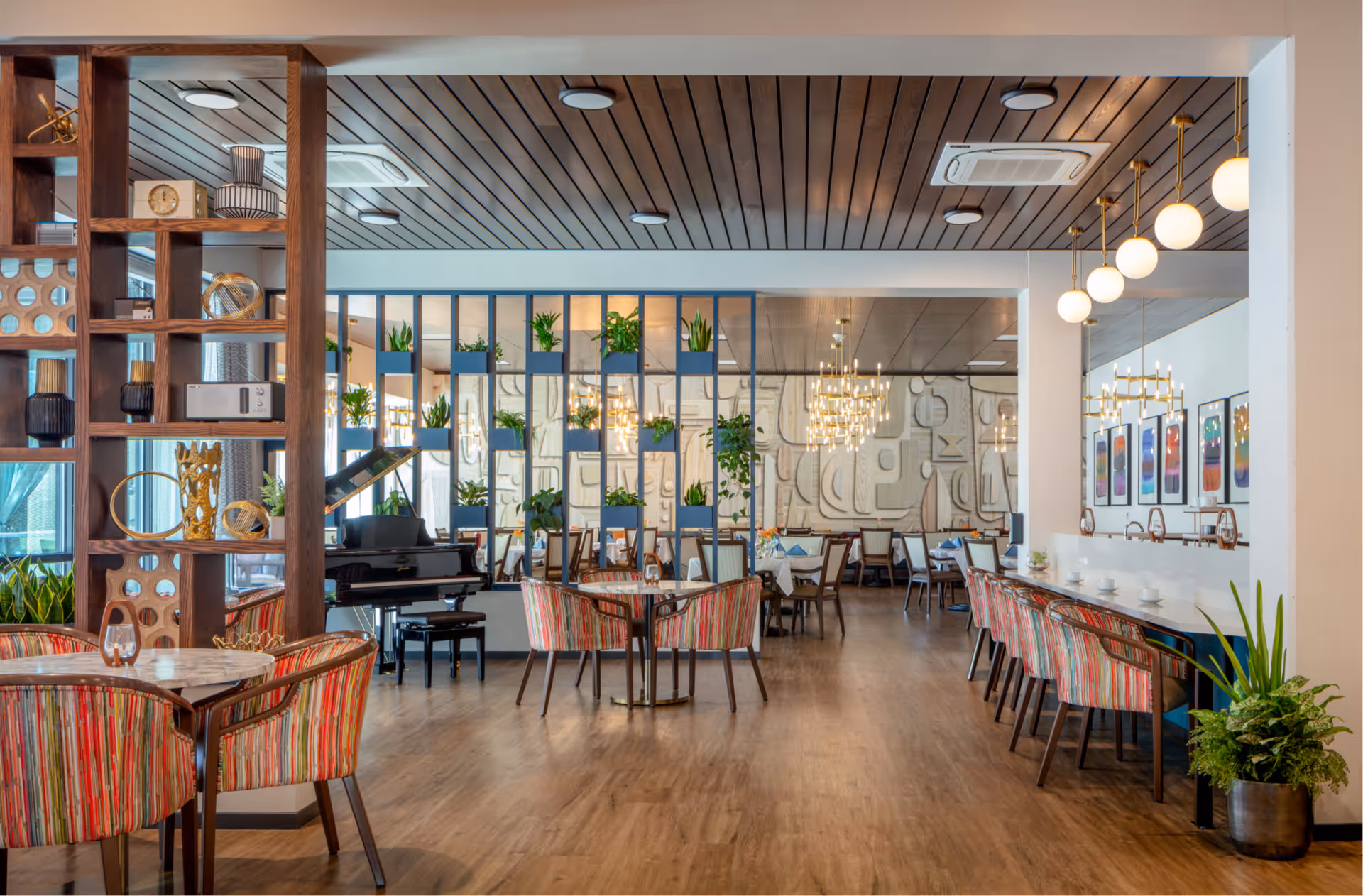 A spacious and modern dining area with wooden flooring and a wooden slatted ceiling. The room features multiple tables with colorful striped chairs, a grand piano, decorative shelving with plants and ornaments, and a wall divider with plants. There are chandeliers and pendant lights hanging from the ceiling, and framed artwork on the walls. The space is bright and inviting with a mix of contemporary and artistic decor.