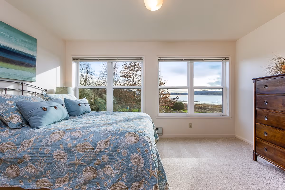 A bright bedroom with a large bed covered in a blue seashell-patterned quilt and matching pillows. There is a wooden dresser on the right side and two large windows showing a view of trees and a body of water outside. A green and blue abstract painting hangs above the bed.