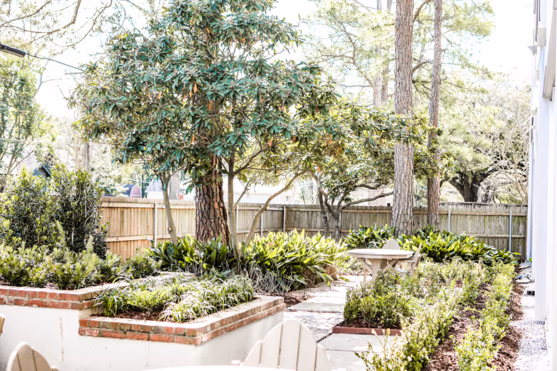 Fenced backyard garden with raised brick planters, trees, pathways, and a small outdoor table and chairs.