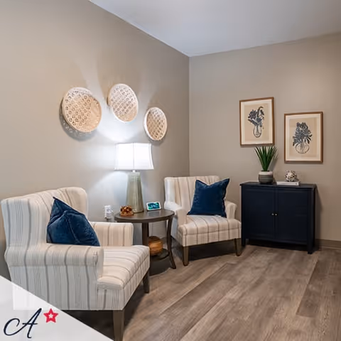 A cozy sitting area with two striped armchairs featuring dark blue throw pillows, a round wooden side table with a lamp and decorative items, three woven wall baskets above the lamp, and a dark blue cabinet with two framed botanical prints and a potted plant on top, all set against beige walls and wood flooring.