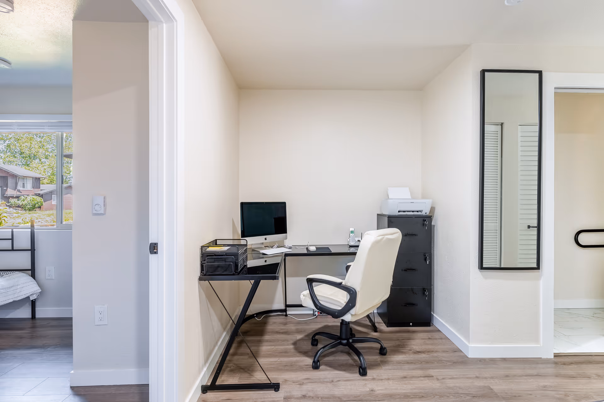 Small home office alcove with a desk, computer, white swivel chair, filing cabinet and wall mirror in a bright apartment.