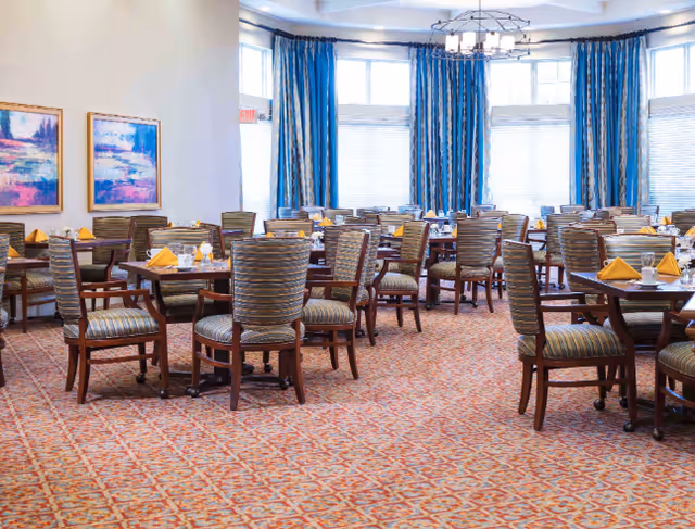 A spacious dining room with multiple wooden tables and striped cushioned chairs. Each table is set with yellow folded napkins, glassware, and white plates. The room features large windows with blue curtains allowing natural light to fill the space. Two colorful abstract paintings hang on the wall to the left, and a modern chandelier hangs from the ceiling.