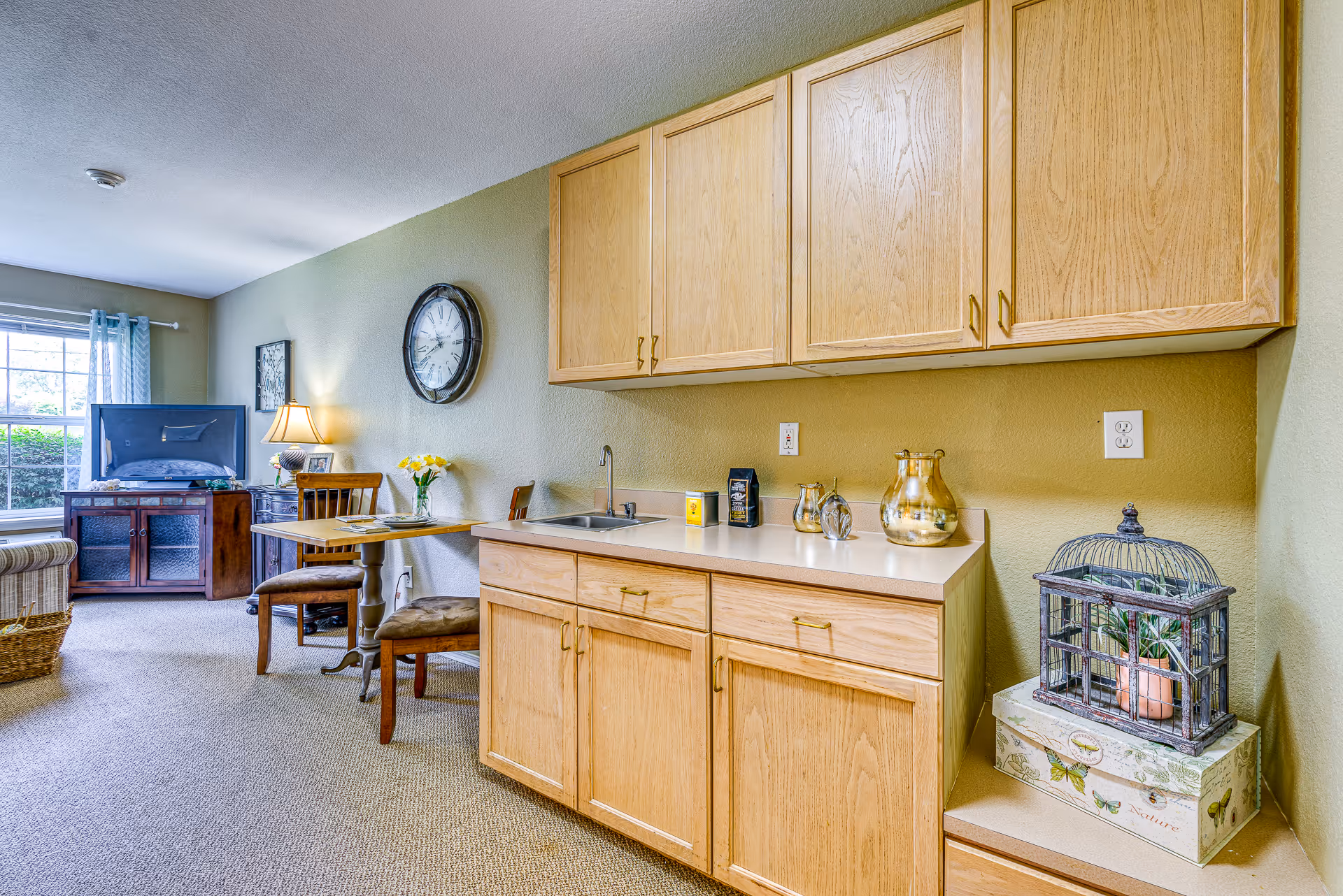 Open studio-style living area with a small kitchenette featuring wooden cabinets and a sink, a dining table, wall clock and a TV by a window.