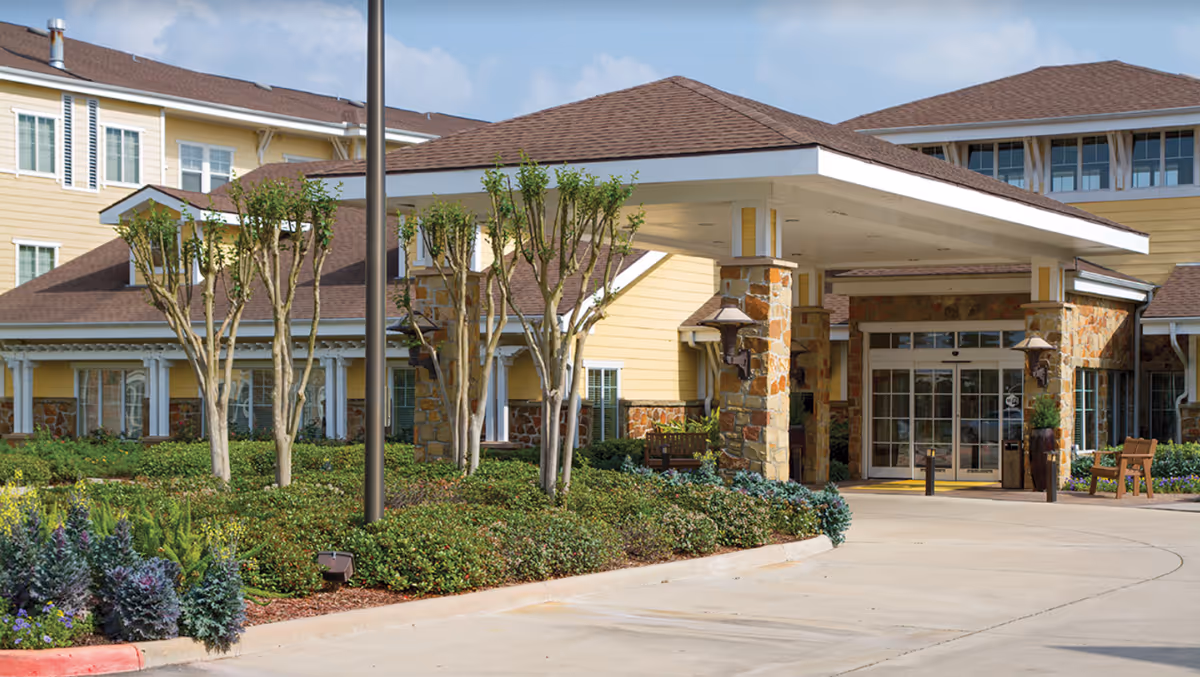 Entrance of The Village at Gleannloch Farms senior living facility showing a covered driveway with stone pillars, landscaped bushes and small trees, and a yellow building with multiple windows under a partly cloudy sky.