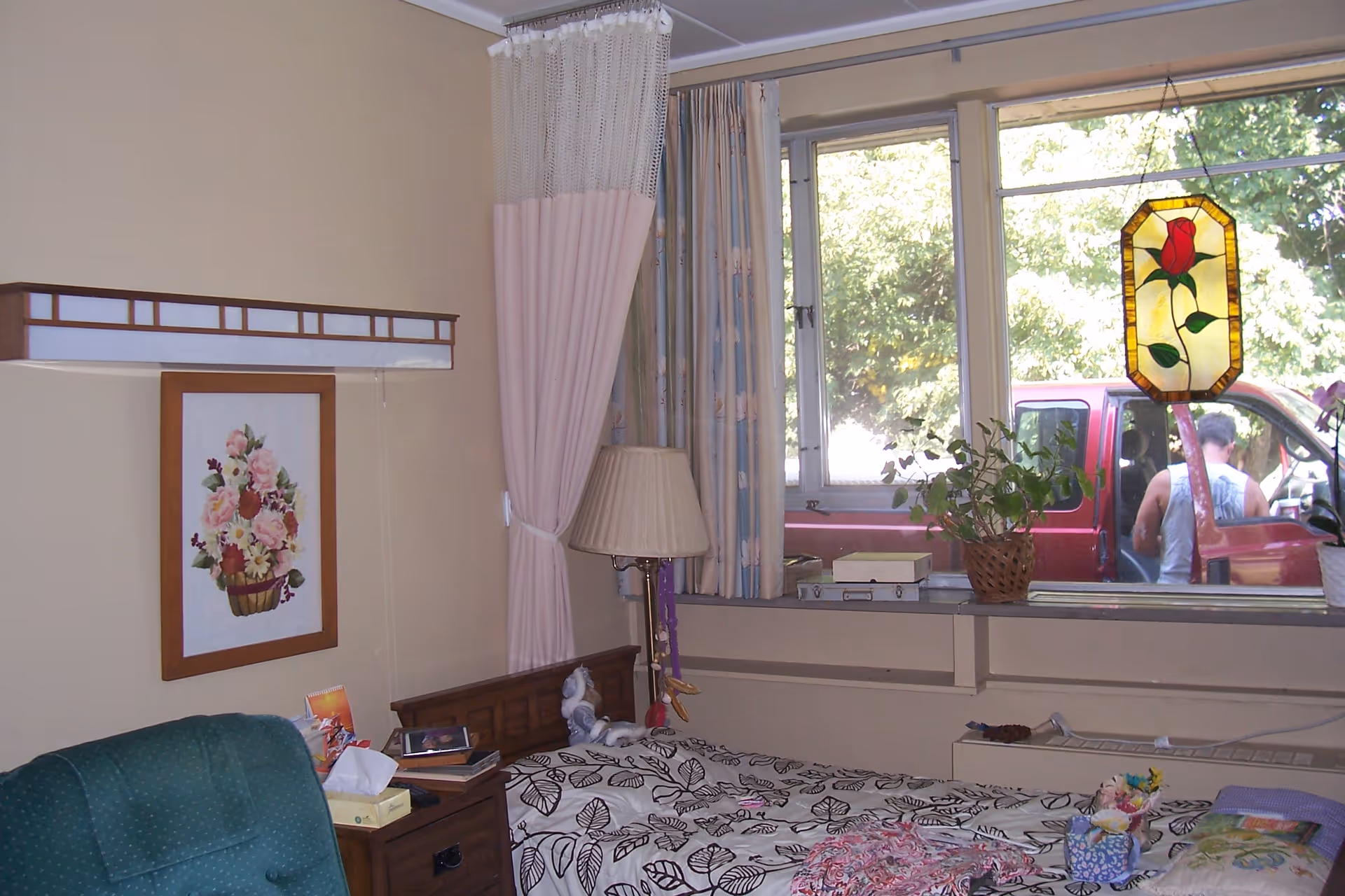 A small bedroom with a single bed, bedside table and lamp, armchair, floral wall art, and a window overlooking a red pickup truck.