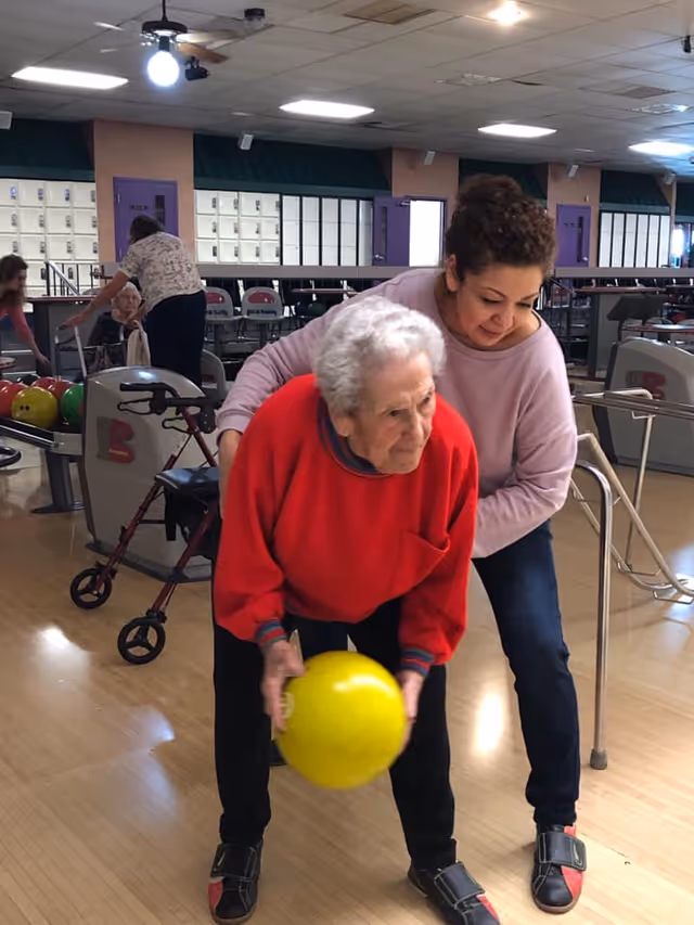 An elderly woman in a red sweater is holding a yellow bowling ball, preparing to bowl, while a younger woman in a light purple top supports her from behind in a bowling alley. In the background, other elderly people and bowling equipment are visible.