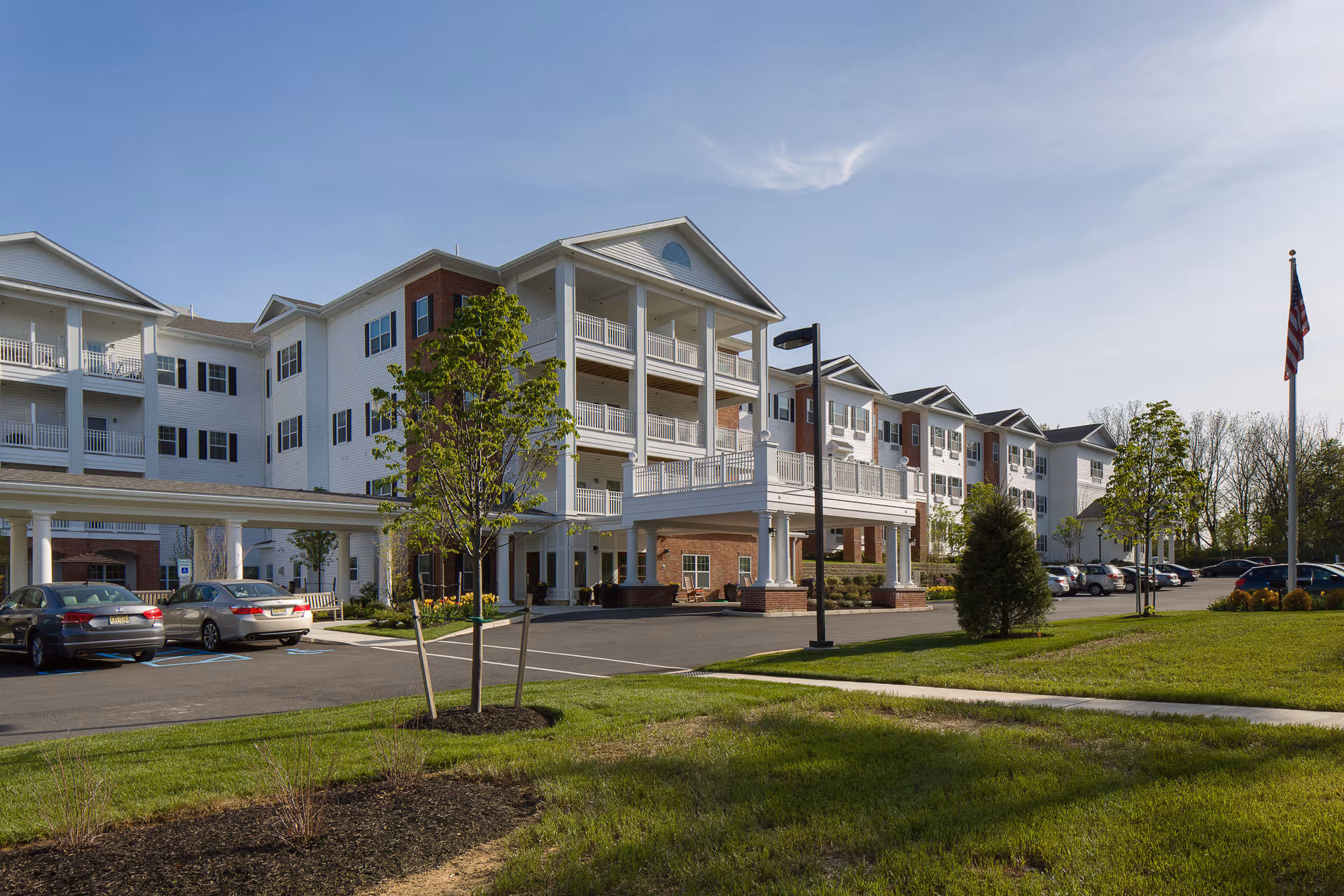 Exterior view of a multi-story senior living facility building with white siding and brick accents, balconies, a covered entrance, parked cars, green lawn, trees, and an American flag on a flagpole under a clear sky.