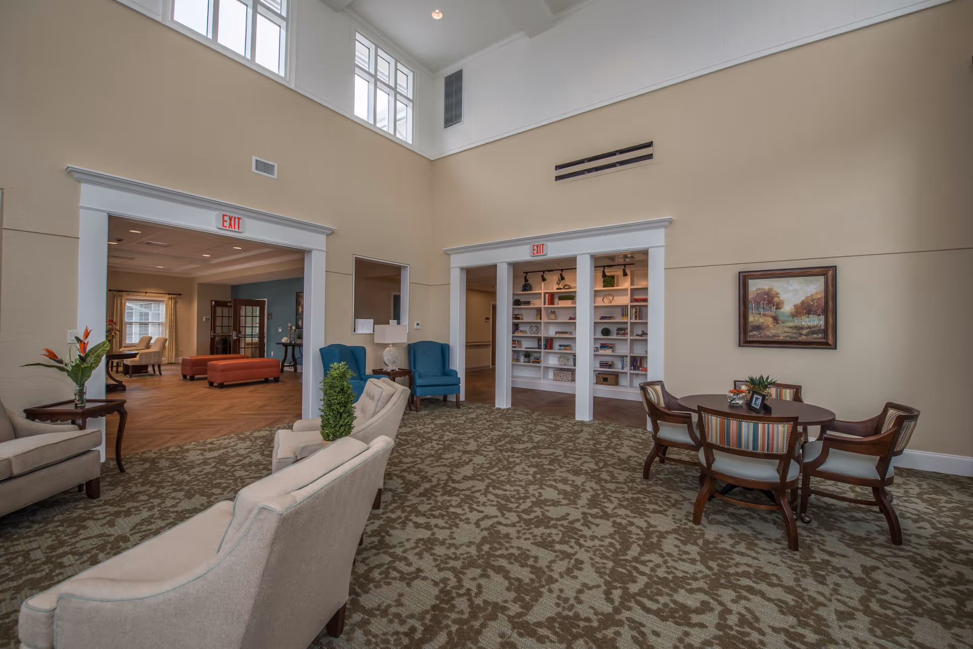 A spacious and well-lit common area in an assisted living facility featuring beige walls and a patterned carpet. There are several seating arrangements including beige armchairs, blue chairs, and a round wooden table with four chairs. The room has high ceilings with windows near the top, and two open doorways leading to other rooms, one with bookshelves and the other with additional seating and a window.