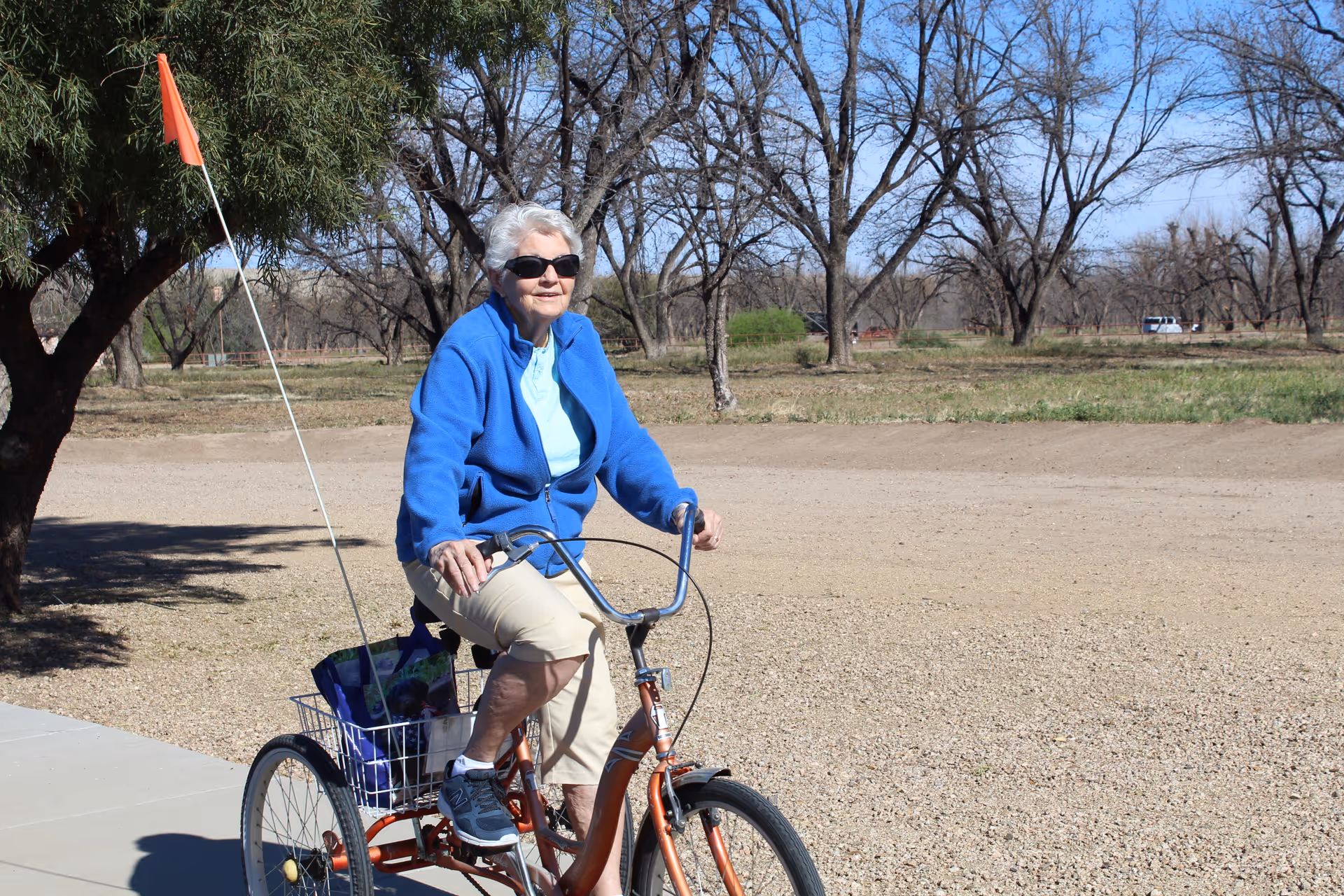 An elderly woman wearing sunglasses, a blue jacket, and beige pants is riding a three-wheeled bicycle on a paved path. The background shows leafless trees and a clear blue sky, indicating a sunny day.