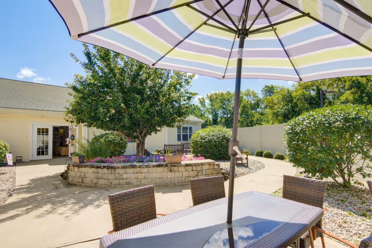 Outdoor patio area at The Atrium at Cardinal Drive featuring a table with a large striped umbrella, surrounded by four wicker chairs. In the background, there is a raised flower bed with colorful flowers and a tree, along with bushes and a curved walkway leading to a building entrance.
