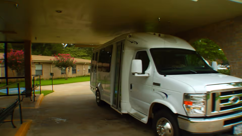 A white shuttle bus parked under a covered driveway area at a nursing center, with a brick building and green lawn with trees in the background.