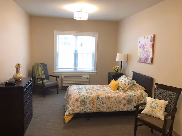 A bedroom with a bed covered in a colorful floral patterned bedspread and two pillows, one yellow and one matching the bedspread. There are two chairs, one with a green blanket draped over it and the other with a decorative pillow. A dresser with a decorative item on top is on the left side of the room. A window with blinds is centered on the far wall above an air conditioning unit. A nightstand with a lamp and a small flower arrangement is next to the bed. A floral painting hangs on the wall above the bed.