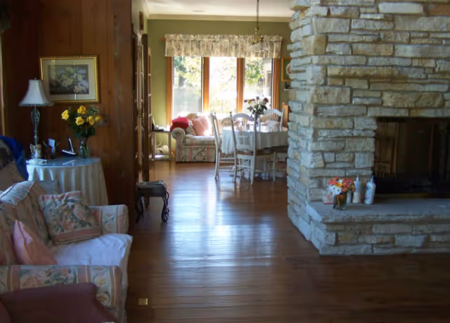A cozy living room with floral-patterned sofas and a side table with a lamp and flowers. The room has wooden floors and a large stone fireplace. In the background, there is a dining area with a table and chairs near large windows letting in natural light.