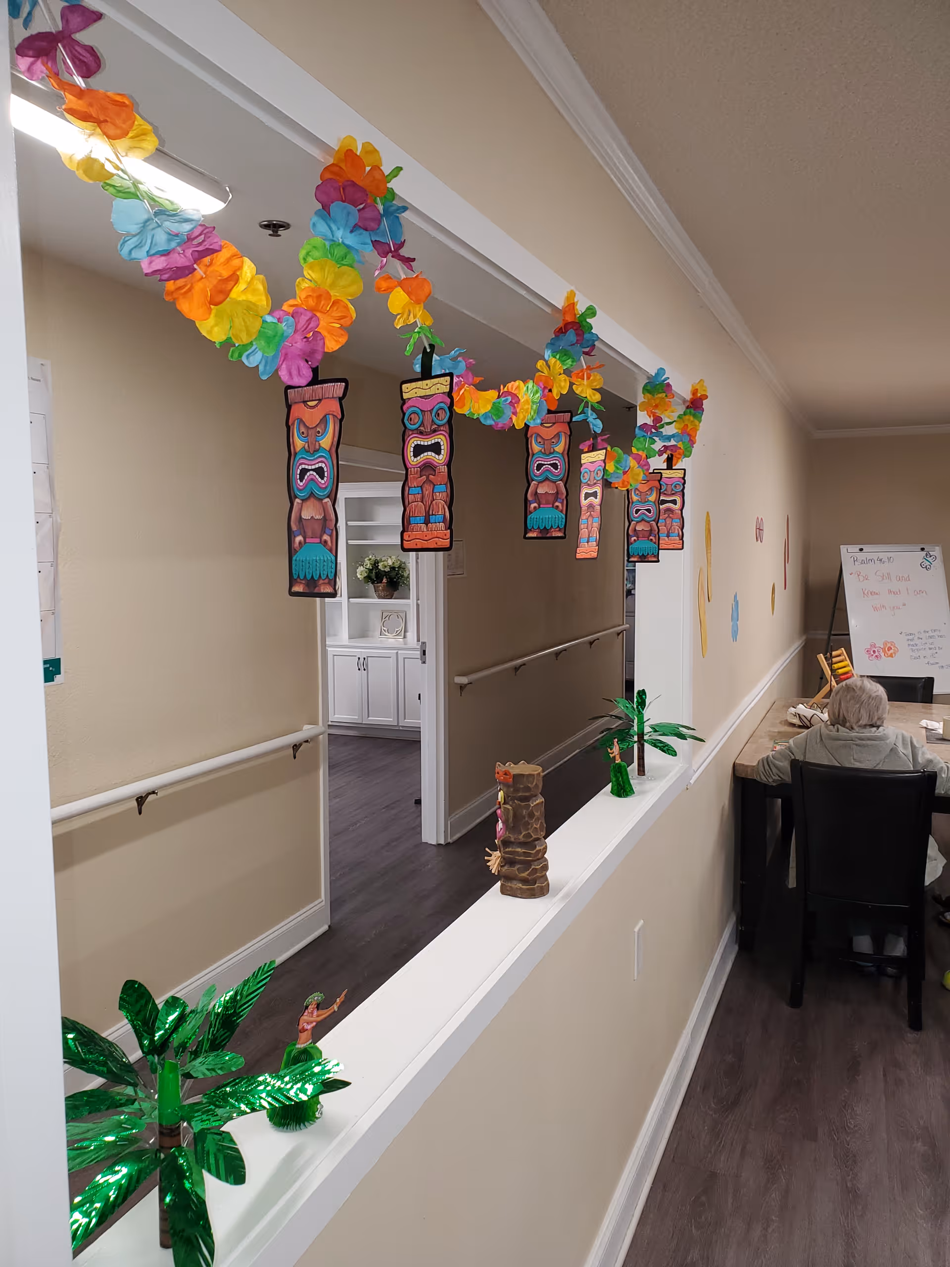 Interior hallway of a senior living facility decorated with colorful Hawaiian-themed garlands and tiki masks hanging from the ceiling. On the ledge, there are small decorative palm trees and tiki figurines. At the end of the hallway, an elderly person is seated at a table with a whiteboard in the background displaying a Bible verse.
