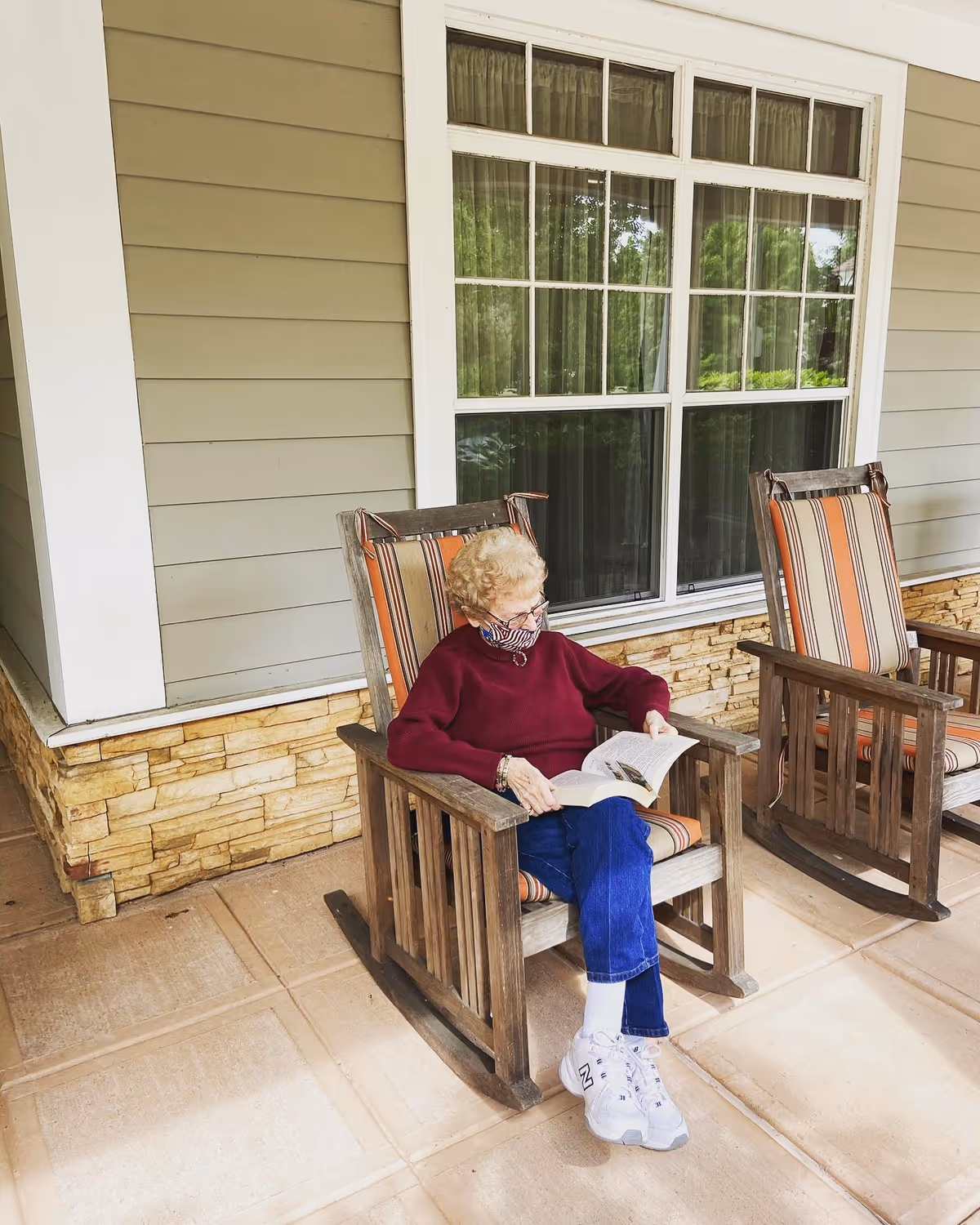 An elderly woman wearing a face mask, a red sweater, blue jeans, and white sneakers is sitting on a wooden rocking chair with striped cushions on a porch. She is reading a book. Another empty rocking chair with similar cushions is next to her. The porch has a stone base and beige siding with a large window behind her.