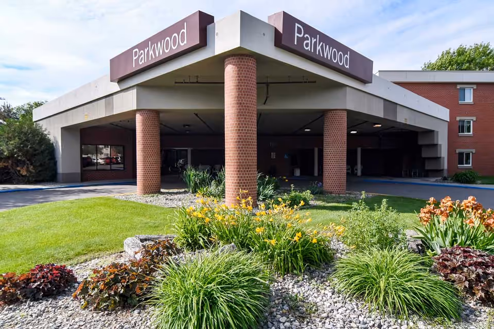 Covered brick-pillared entrance of the Parkwood senior living building with landscaped flower beds and lawn in front.