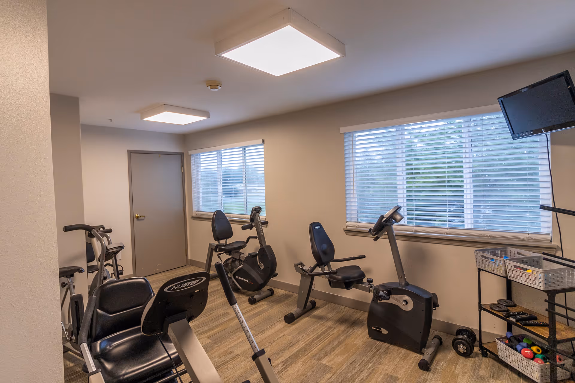 A small exercise room with stationary bikes and other fitness equipment. The room has two large windows with blinds, light-colored walls, and wood-patterned flooring. There is a wall-mounted TV in the corner and a shelving unit holding various small weights and exercise accessories.