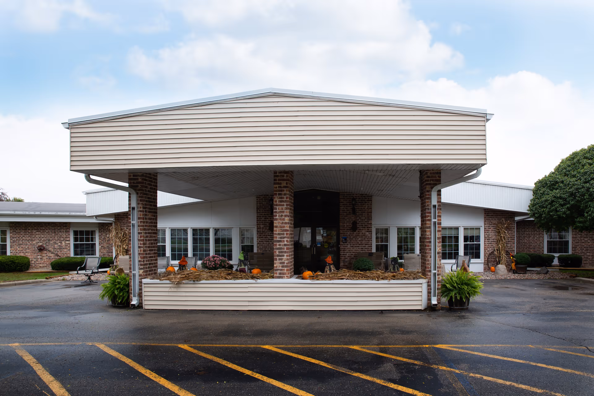 Front entrance of a brick senior living facility with a covered drop-off, columns, and autumn decorations including pumpkins and corn stalks.