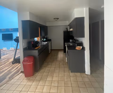Narrow galley kitchen with dark cabinets, tiled floor, appliances, and a red trash bin opening into a room with a treadmill and wall-mounted TV.