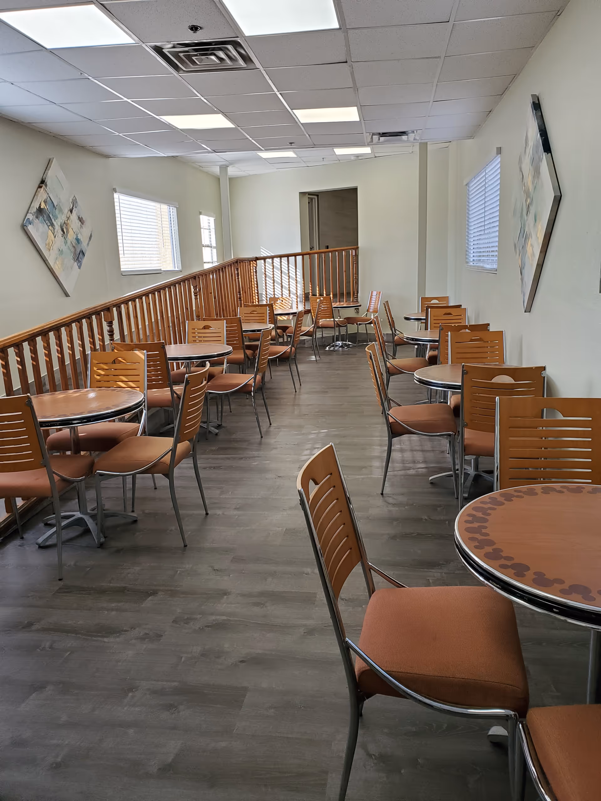 A dining area with multiple round tables and orange cushioned chairs arranged neatly along a wooden railing. The room has light-colored walls with two abstract paintings, windows with blinds, and a ceiling with fluorescent lights.