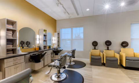 Bright salon-style room with styling chairs, shampoo sinks, mirrors, shelving with towels, and hooded dryers.