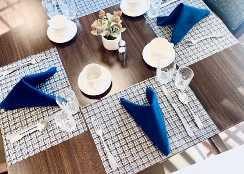 Wood dining table set for four with blue folded napkins, cups, glasses, silverware, patterned placemats and a small floral centerpiece.