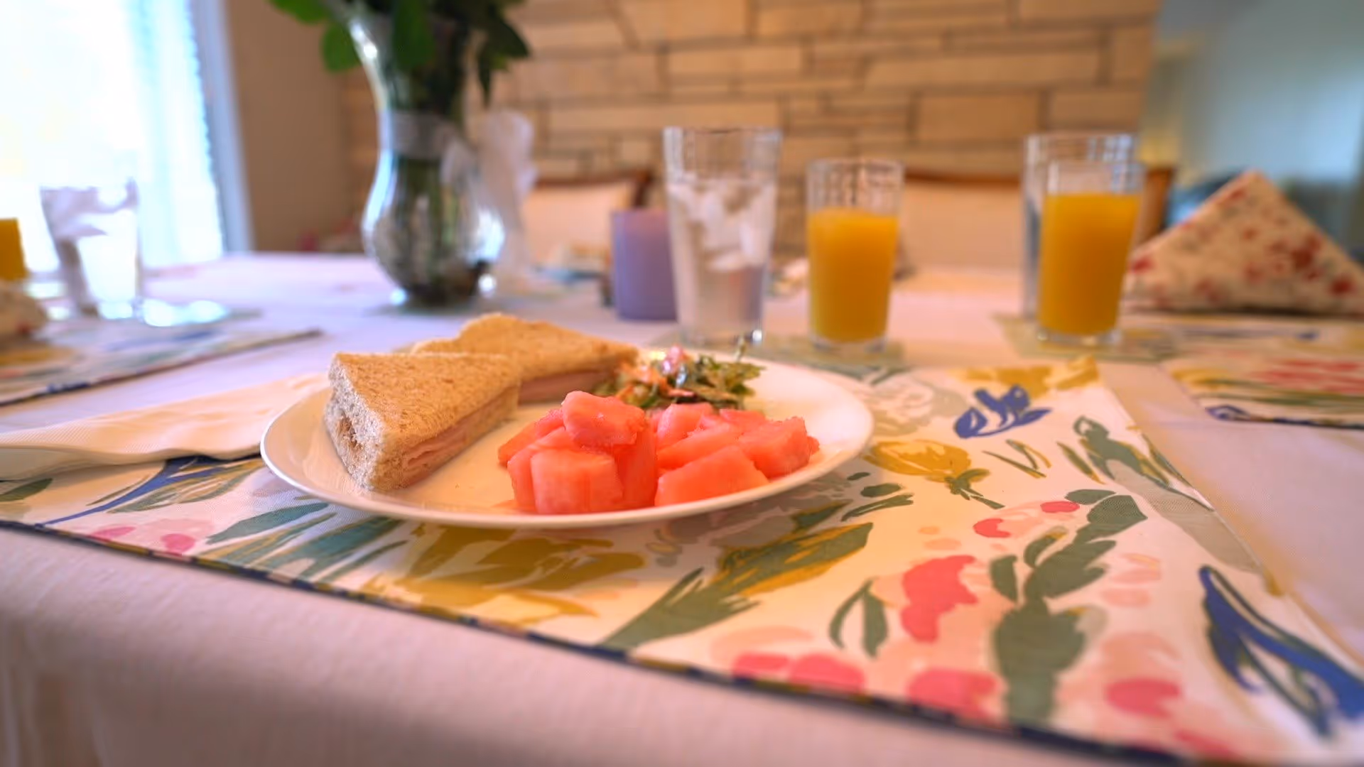 A close-up of a dining table set with a plate containing a sandwich, watermelon chunks, and a small salad. There are glasses of water and orange juice on the table, which is covered with a floral-patterned placemat and a white tablecloth. The background shows a stone wall and cushioned chairs.
