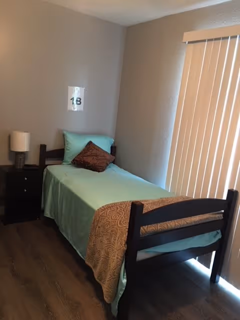 A small bedroom with a single bed covered in light blue bedding and a brown decorative pillow. There is a wooden nightstand with a lamp next to the bed. Vertical blinds cover the window, and a sign with the number 18 is taped to the wall above the bed.