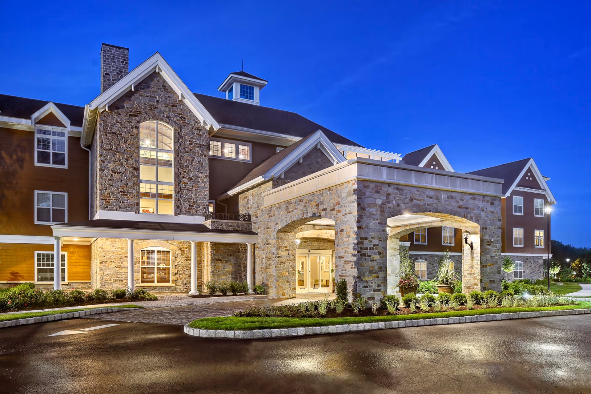 Exterior view of a senior living facility building at dusk with stone and wood siding, large windows, and a covered entrance with stone pillars and landscaping.